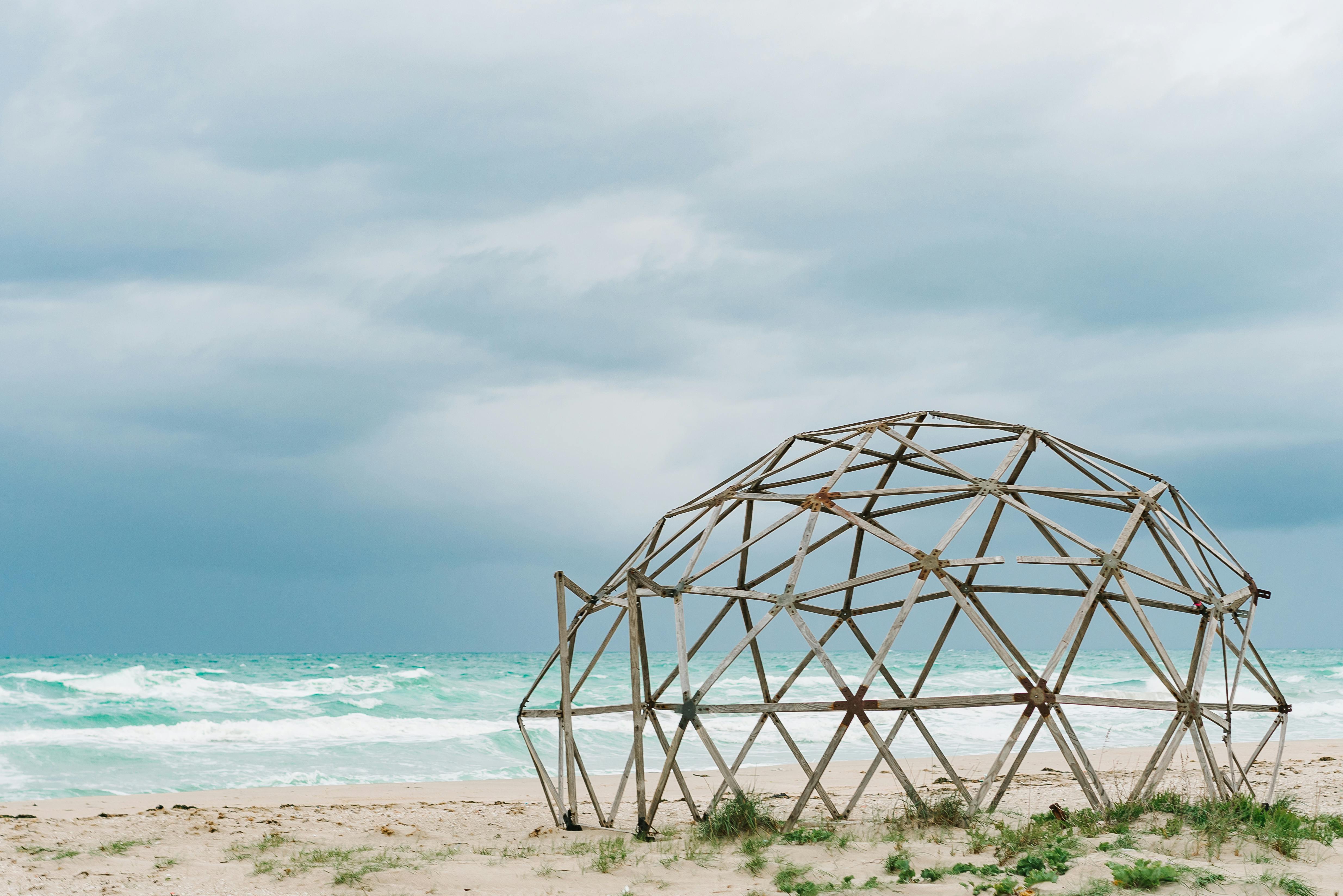 A rustic geodesic dome structure on a sandy beach with a cloudy sky overhead.