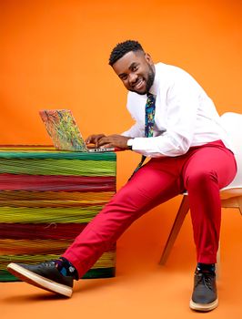 Businessman in bright attire typing on laptop against orange backdrop, exuding positivity.