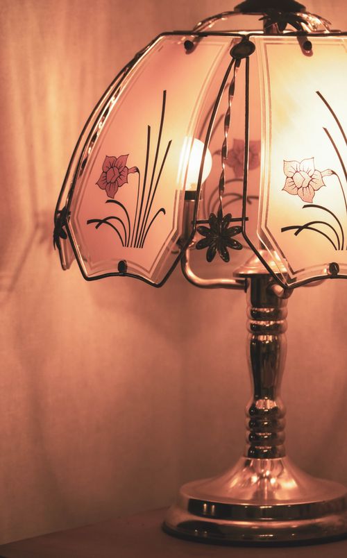 Free A close-up of a vintage table lamp with a floral patterned glass shade radiating a warm glow. Stock Photo