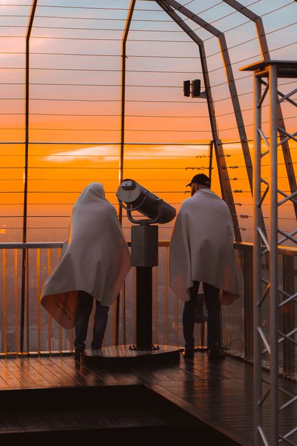 Two people covered with blankets enjoy the sunset from an observation deck in the city.