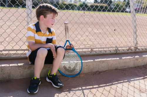Free A young boy sits with a tennis racket by a chain-link fence on a sunny day, embodying youthful leisure. Stock Photo