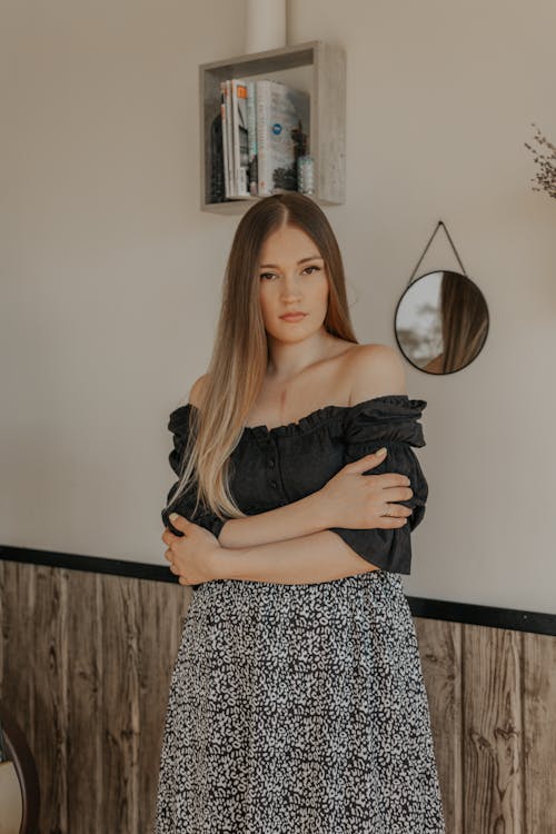 Trendy young female in crop top and ornamental skirt looking at camera with folded arms in house