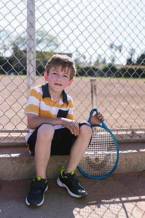 Free A smiling young boy in casual attire sitting outside with a tennis racket by a chain-link fence. Stock Photo