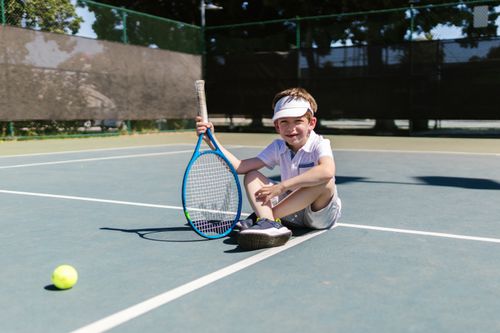 Free A cheerful boy sitting on a tennis court with a racket and ball, enjoying a sunny day. Stock Photo