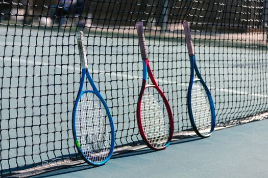 Three colorful tennis rackets leaning against a net on an outdoor tennis court.