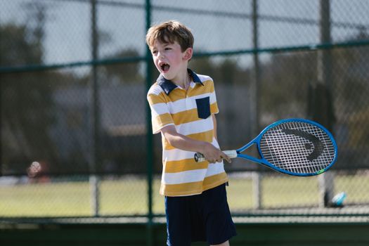 A young boy enthusiastically swings a tennis racket on an outdoor court.