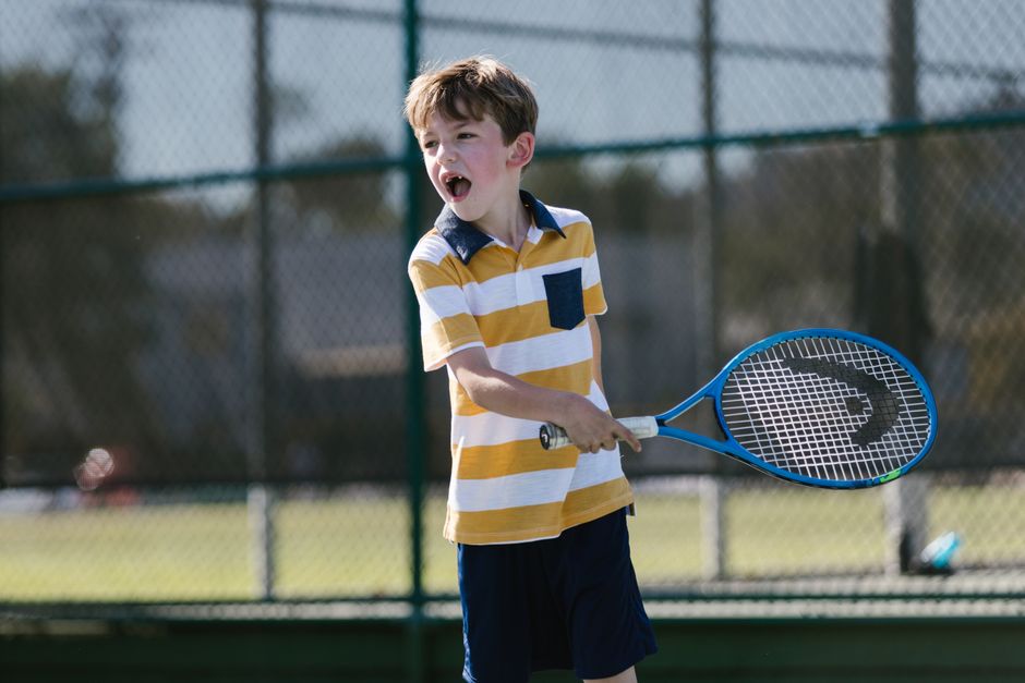 A young boy enthusiastically swings a tennis racket on an outdoor court.