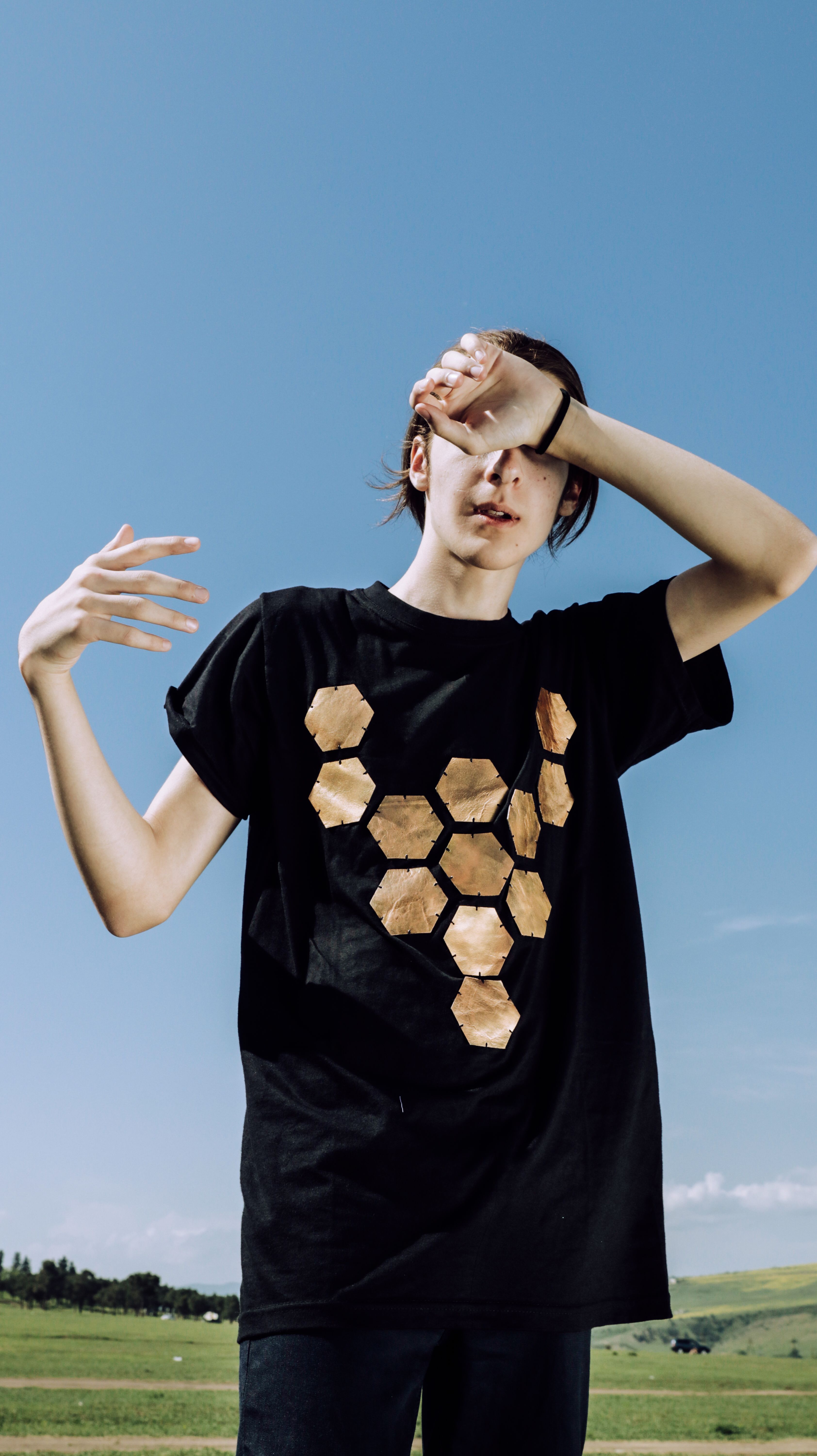 Person in black t-shirt with hexagonal design outdoors shielding eyes from sunlight.