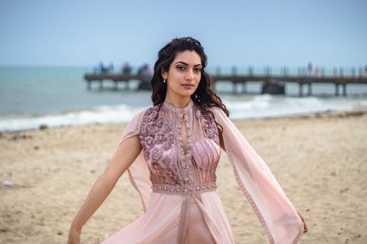 Woman posing gracefully on a beach in a detailed pink dress, showcasing elegance and style.