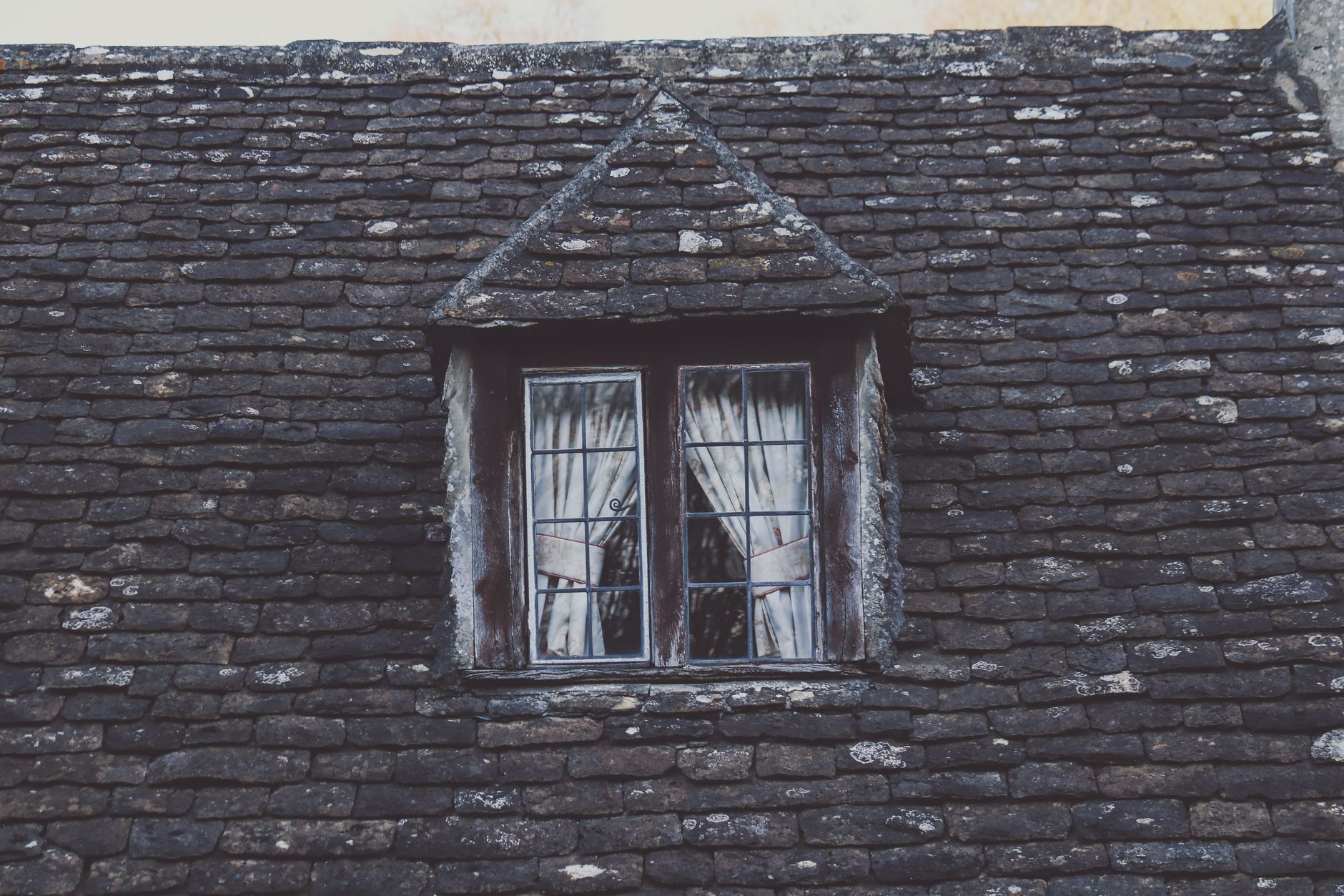 窗簾訂製老屋窗型不正會加價嗎?常見施工費用全解析 A close-up of a rustic stone roof featuring a vintage dormer window with curtains, showcasing architectural details.