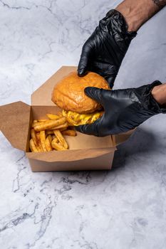 Delicious burger and seasoned fries in a cardboard takeout box, held with gloved hands.