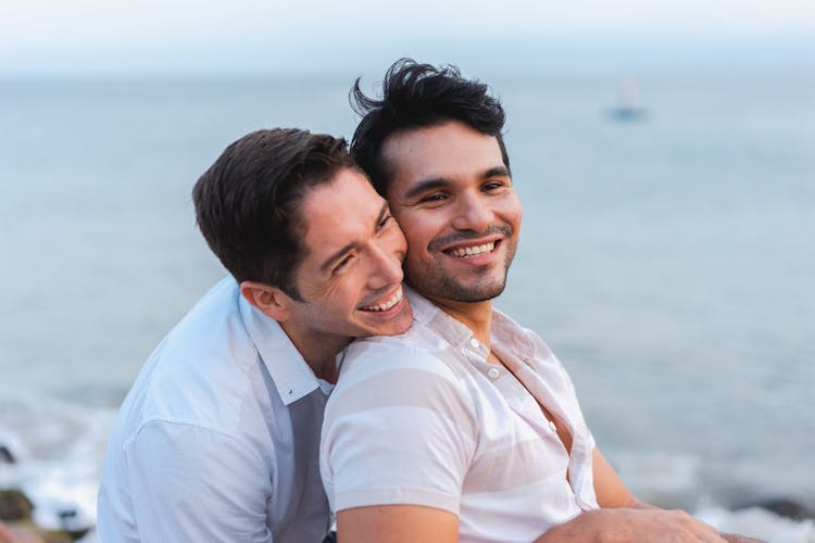 Gay Couple Sitting On A Seashore, Hugging And Smiling 