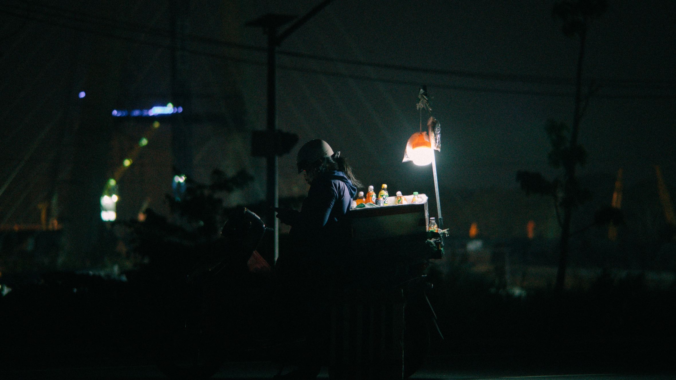 A street vendor sells bottled drinks from an illuminated cart on a Vietnam street at night.