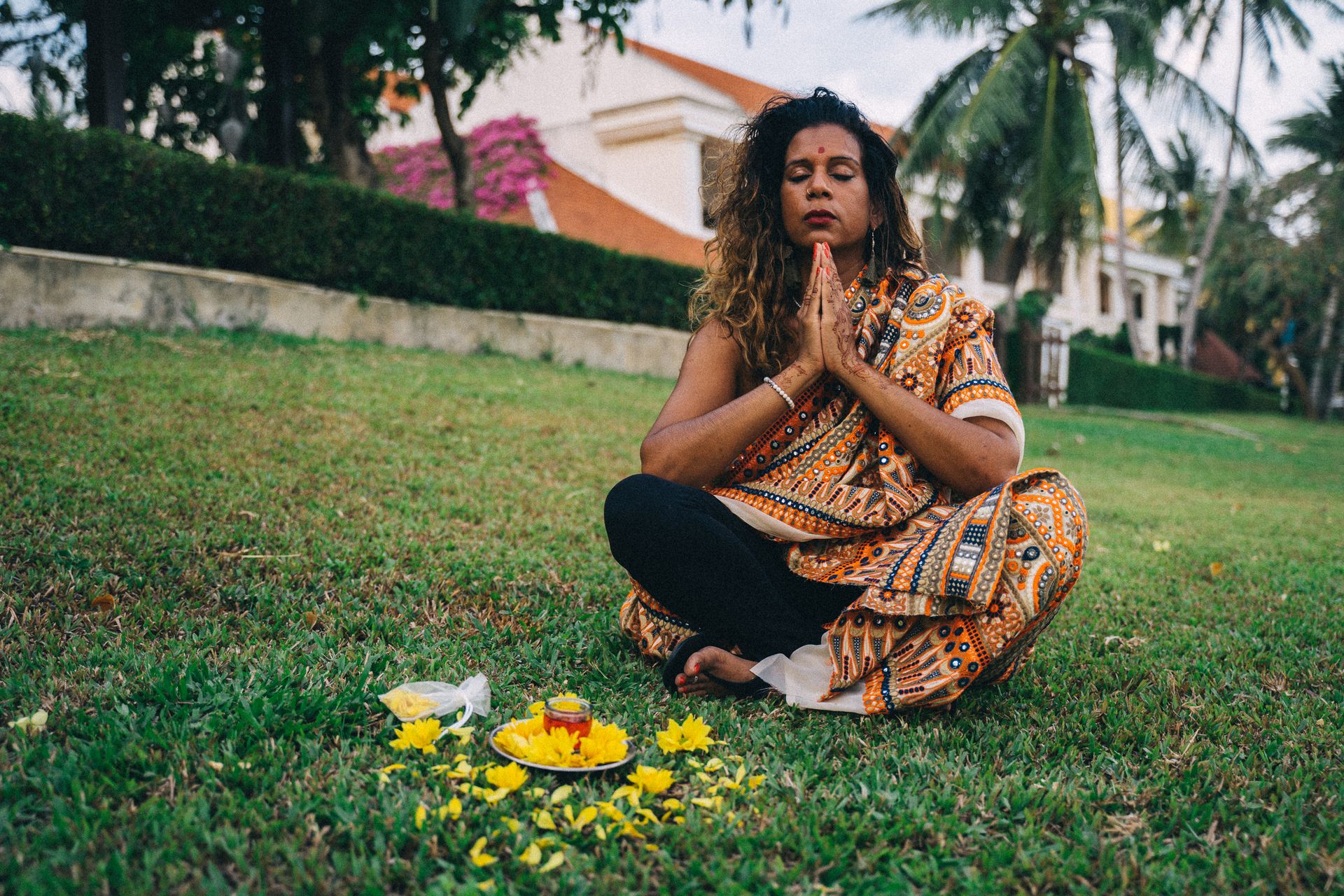 A woman in traditional Indian dress meditates outdoors with prayer hands and yellow flowers.