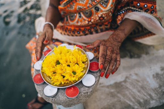 Close-up of a woman's henna-decorated hands holding a plate of candles and yellow flowers for Diwali.