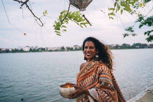Woman in a Traditional Saree Dress Holding a Bowl and Standing by a Body of Water 