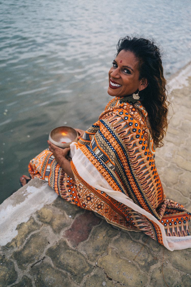 A Woman Wearing Saree Sitting Near The Body Of Water While Holding A Bowl With Lighted Candle