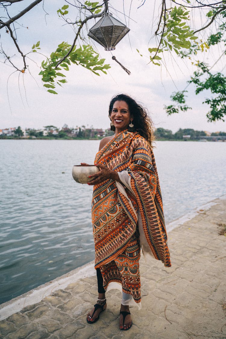 A Woman In Printed Saree Standing Near The Body Of Water While Holding A Bowl