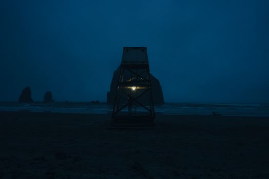A lit lifeguard tower stands against the misty Haystack Rock at Cannon Beach during nighttime.