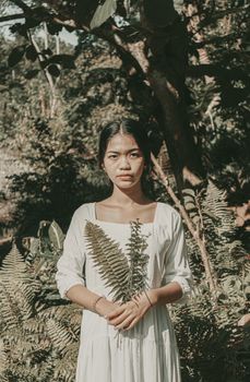 Asian woman in a white dress holds fern leaves in a Bali forest, for a nature portrait.