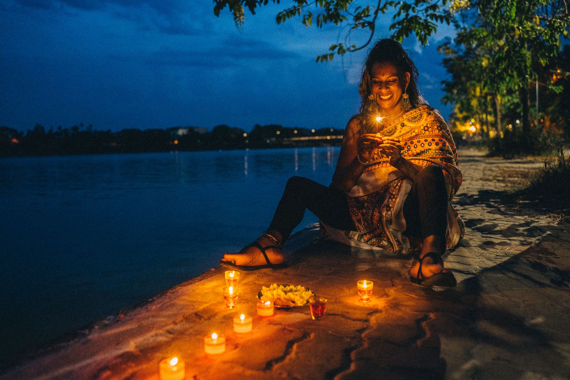 A joyful Indian woman celebrates Diwali with candles and sparklers by the river at night.