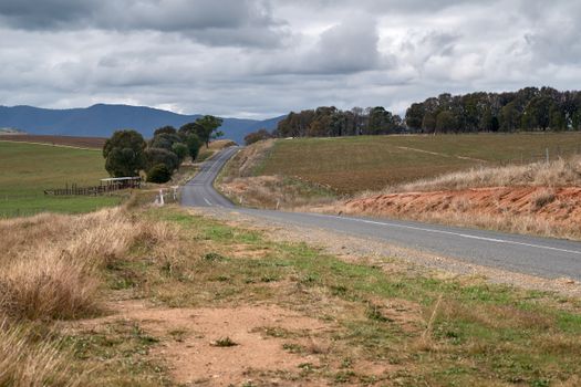 A picturesque rural road winds through the rolling hills of the Australian countryside under a cloudy sky.