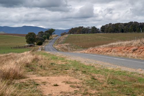 Free A picturesque rural road winds through the rolling hills of the Australian countryside under a cloudy sky. Stock Photo