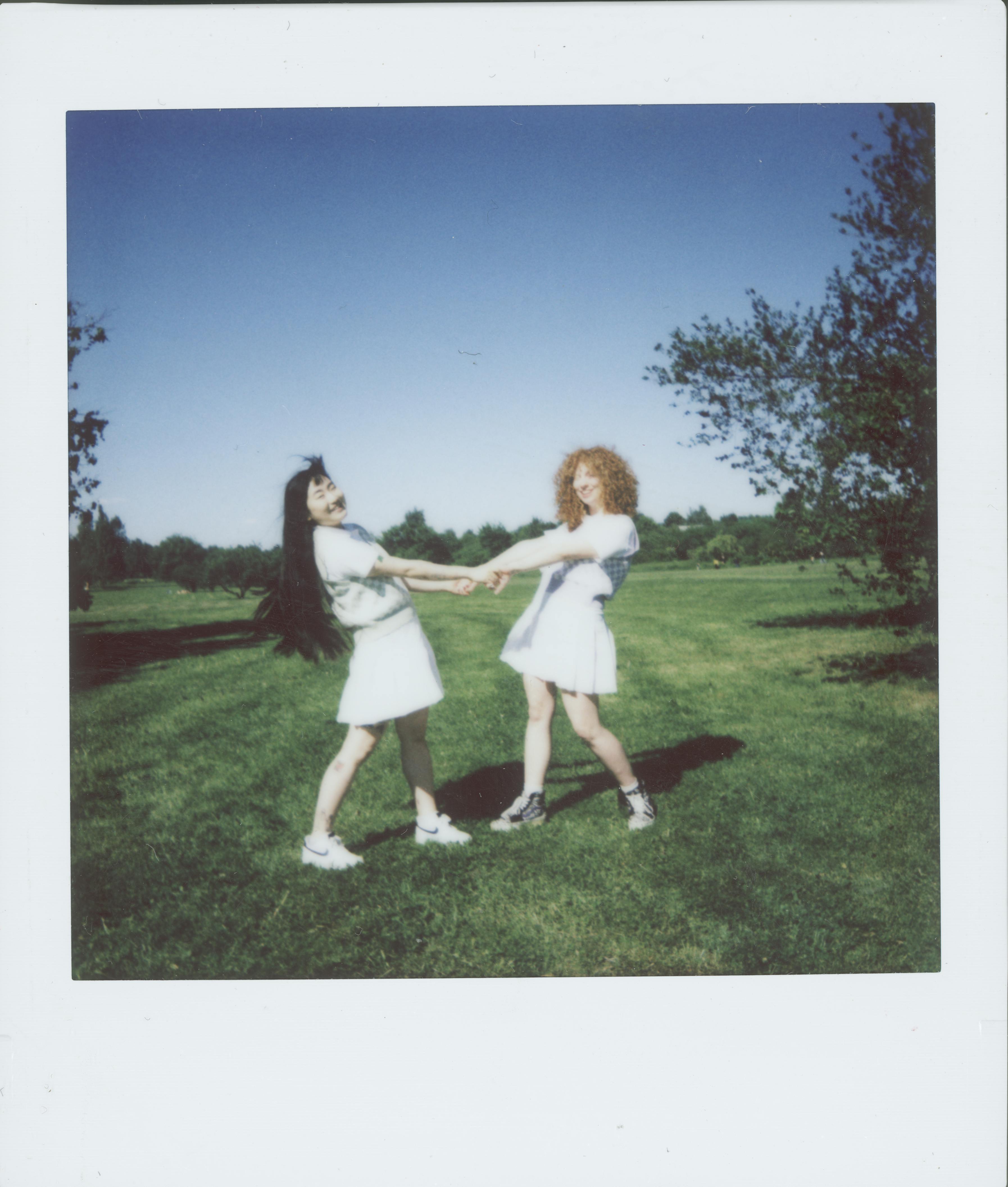 Two women in school uniforms holding hands and smiling in a sunny park, captured in a Polaroid style.