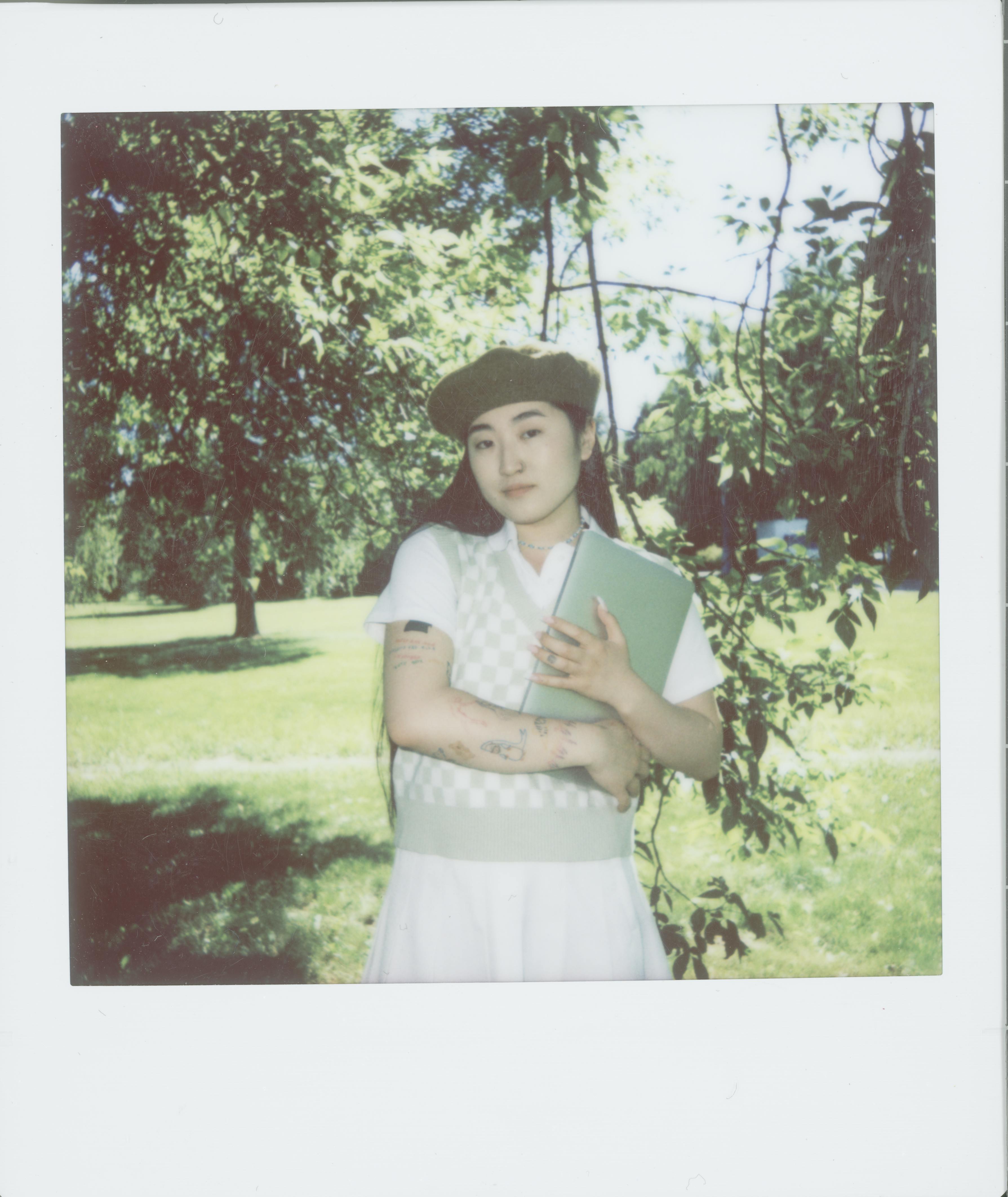 A young woman holding a book stands in a sunny park, surrounded by trees and greenery.