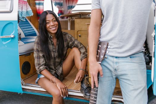 A cheerful woman sits in a campervan door while another person holds a guitar nearby.