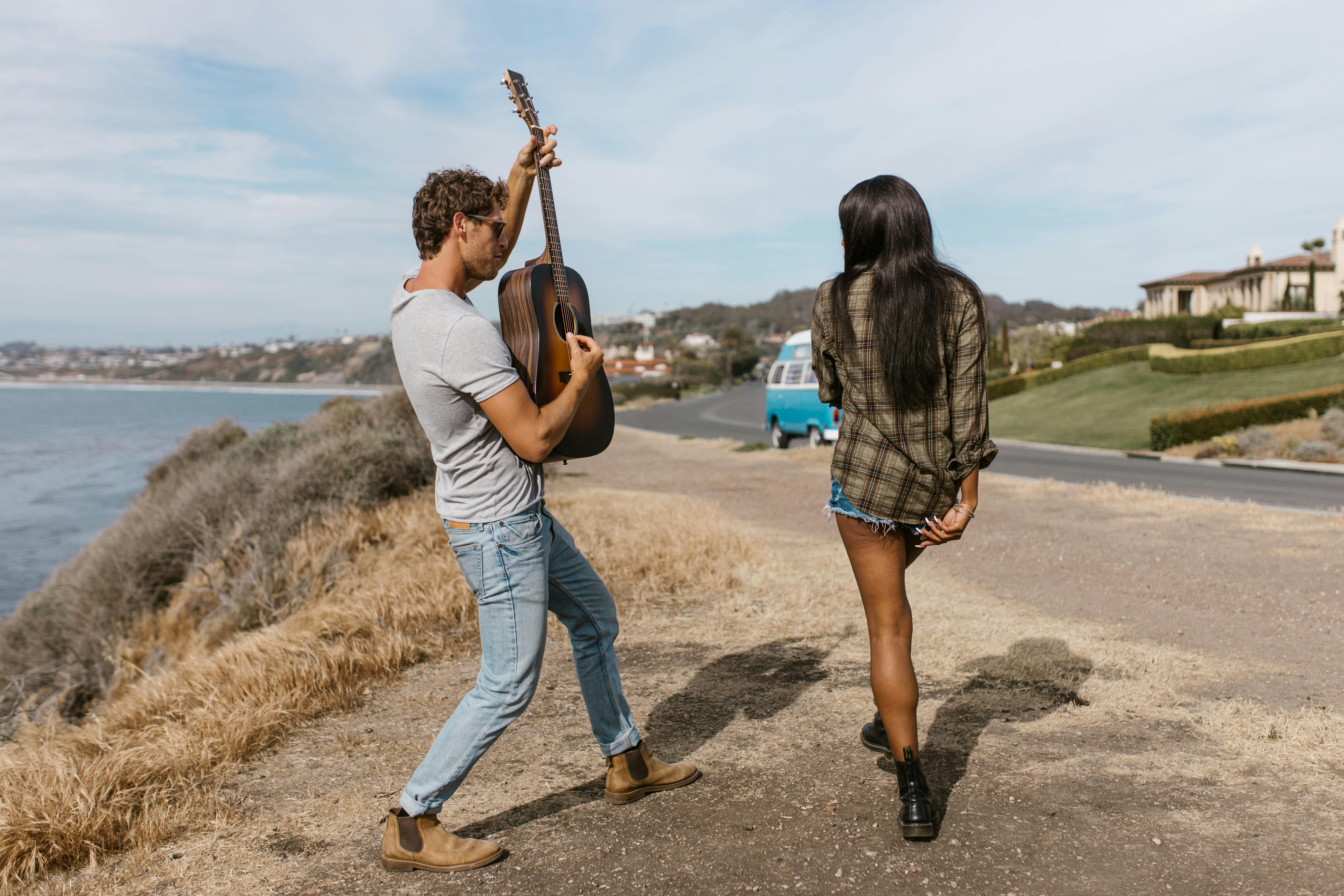A relaxed young couple on a coastal road trip, with the man playing guitar and a VW van in the background.