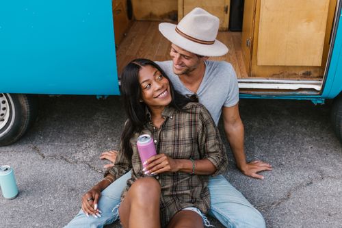 Free A joyful couple sitting together by a blue campervan, sharing a drink and enjoying a sunny day. Stock Photo