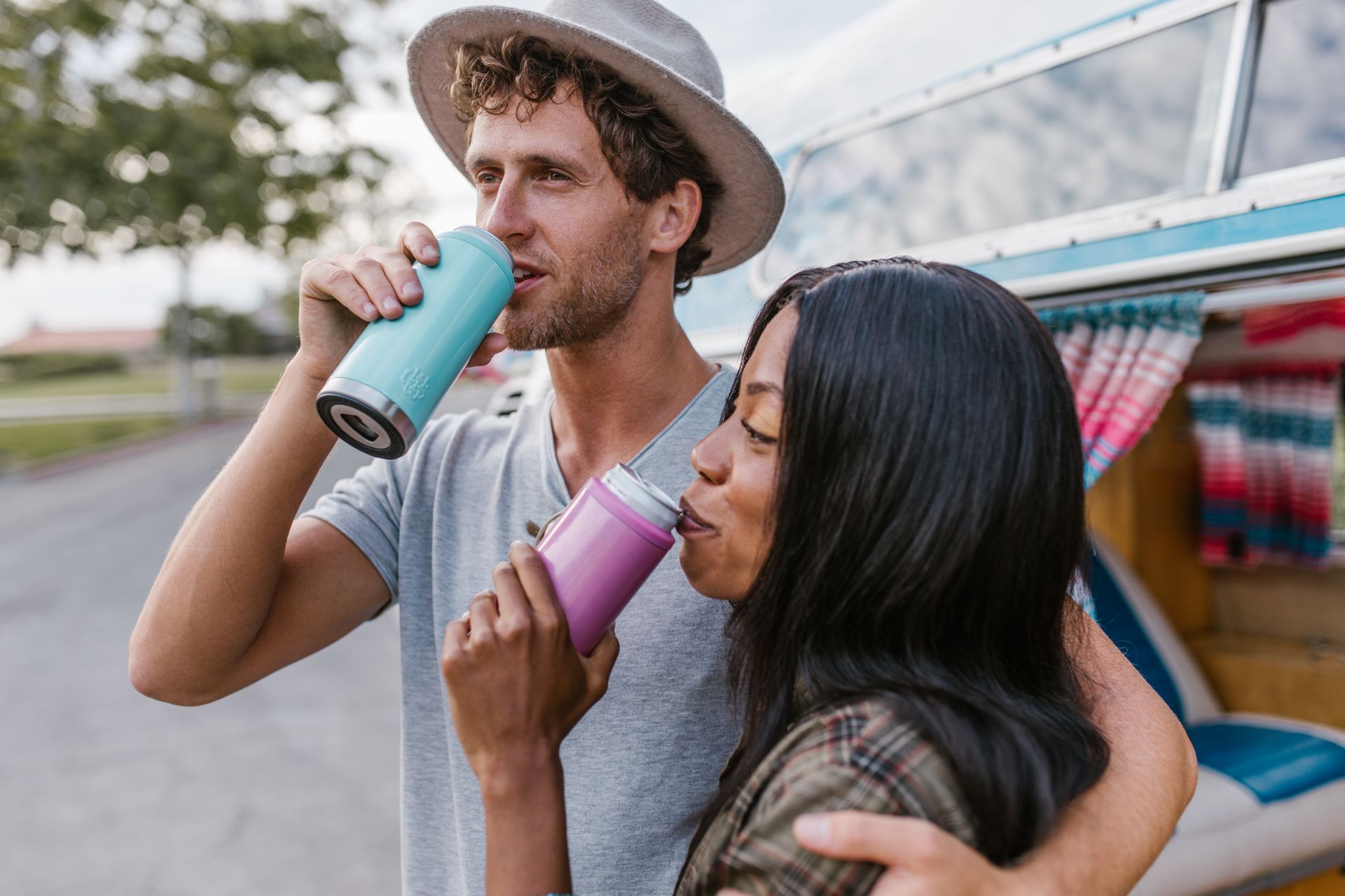 A diverse couple drinking from tumblers outside a van, embracing and enjoying the moment.