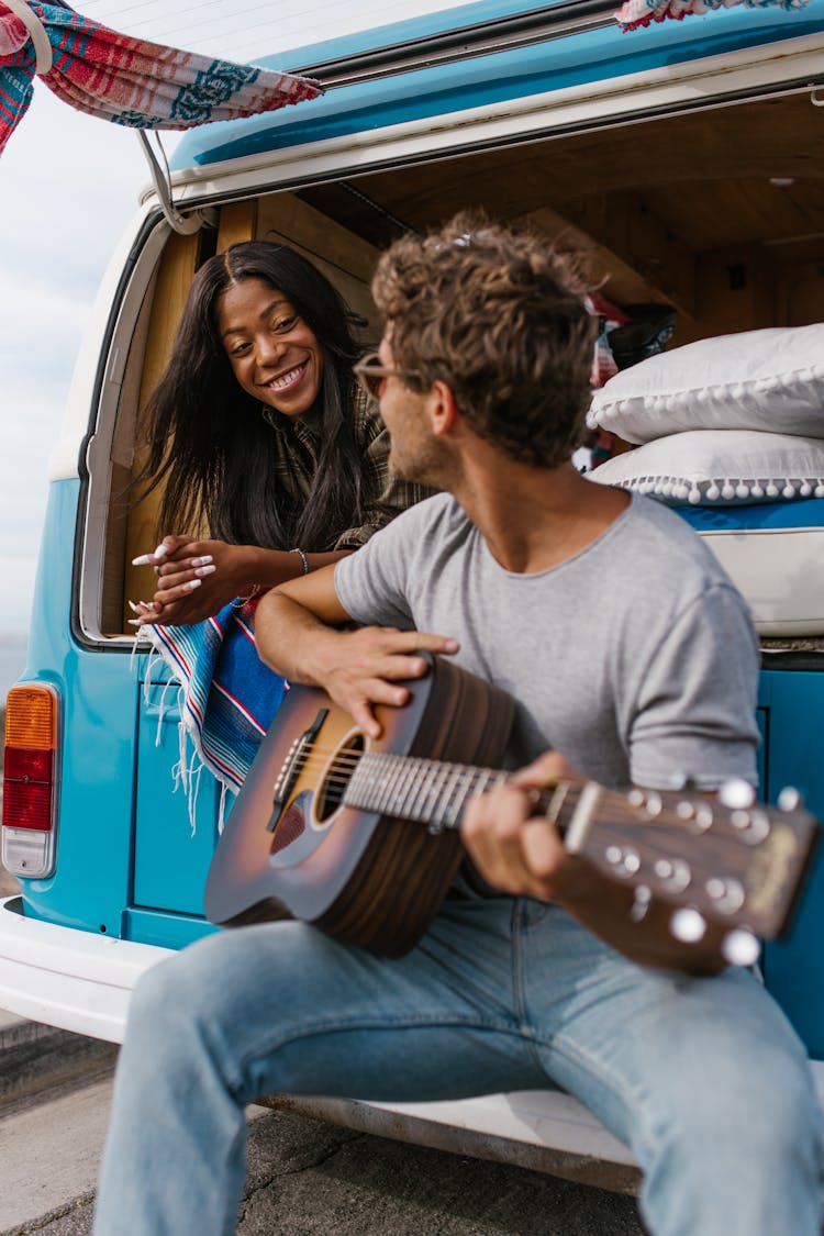 A Woman Looking At A Man With A Guitar Sitting At The Back Of A Van