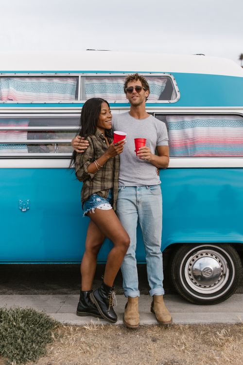 Free Happy interracial couple with red cups by a vintage blue van enjoying a sunny day. Stock Photo