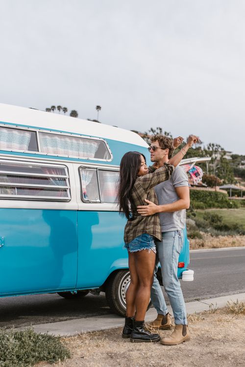 Free Happy couple dancing beside a retro blue van on a sunny day, embracing carefree summer vibes. Stock Photo