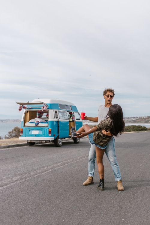 Free A happy couple dances next to a vintage van parked on a scenic road, evoking freedom and adventure. Stock Photo