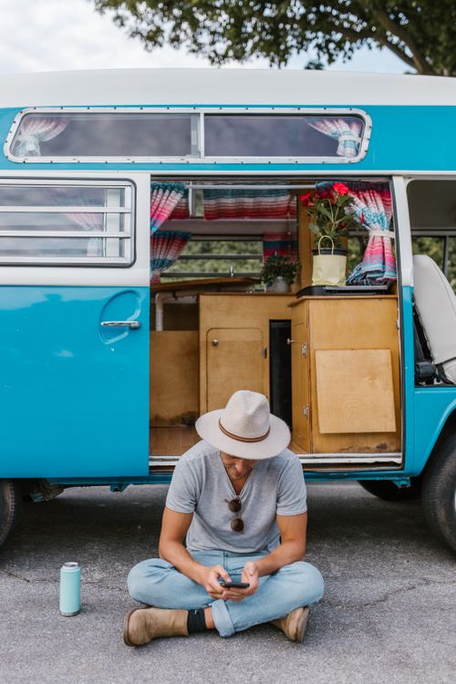Free Man sitting by colorful camper van, fully engaged in outdoor leisure travel. Stock Photo