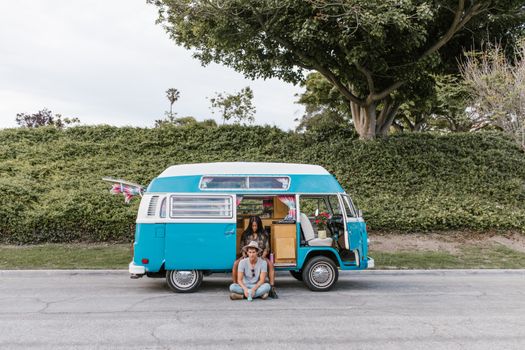 A young couple relaxes by their vintage blue camper van parked on a street in the summer.