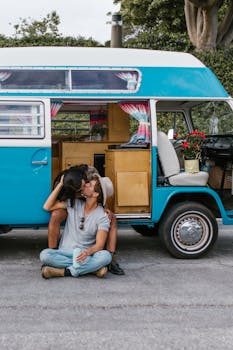 A couple shares a kiss while seated by a retro campervan in an outdoor setting.