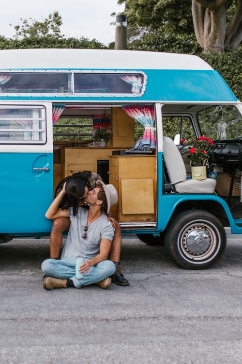 Free A couple shares a kiss while seated by a retro campervan in an outdoor setting. Stock Photo