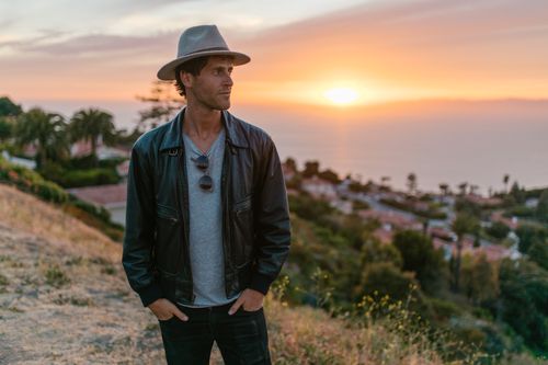 Free A man in a leather jacket and hat stands outdoors at sunset, overlooking a coastal view. Stock Photo