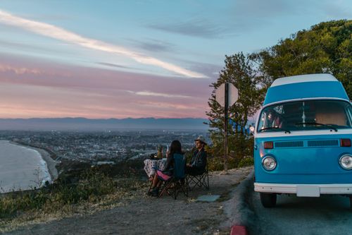Free A couple enjoys a picnic at sunset beside a vintage van with a breathtaking coastal city view. Stock Photo