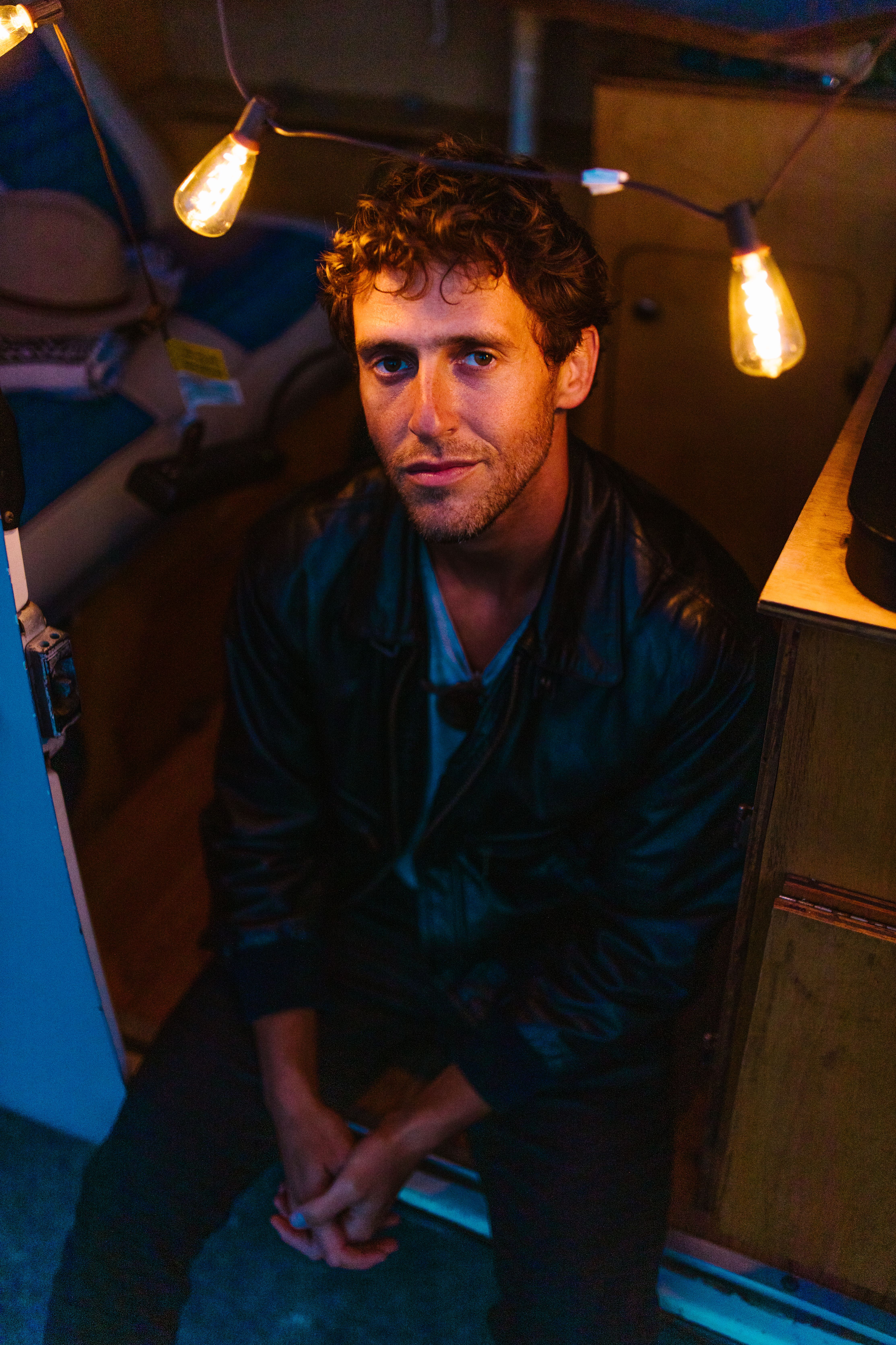 Dramatic portrait of a man with curly hair under warm ambient string lights.