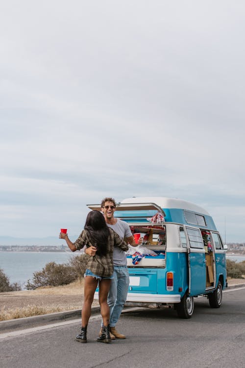Couple Dancing with Drinks in Their Hand next to a Campervan Parked on the Shore 
