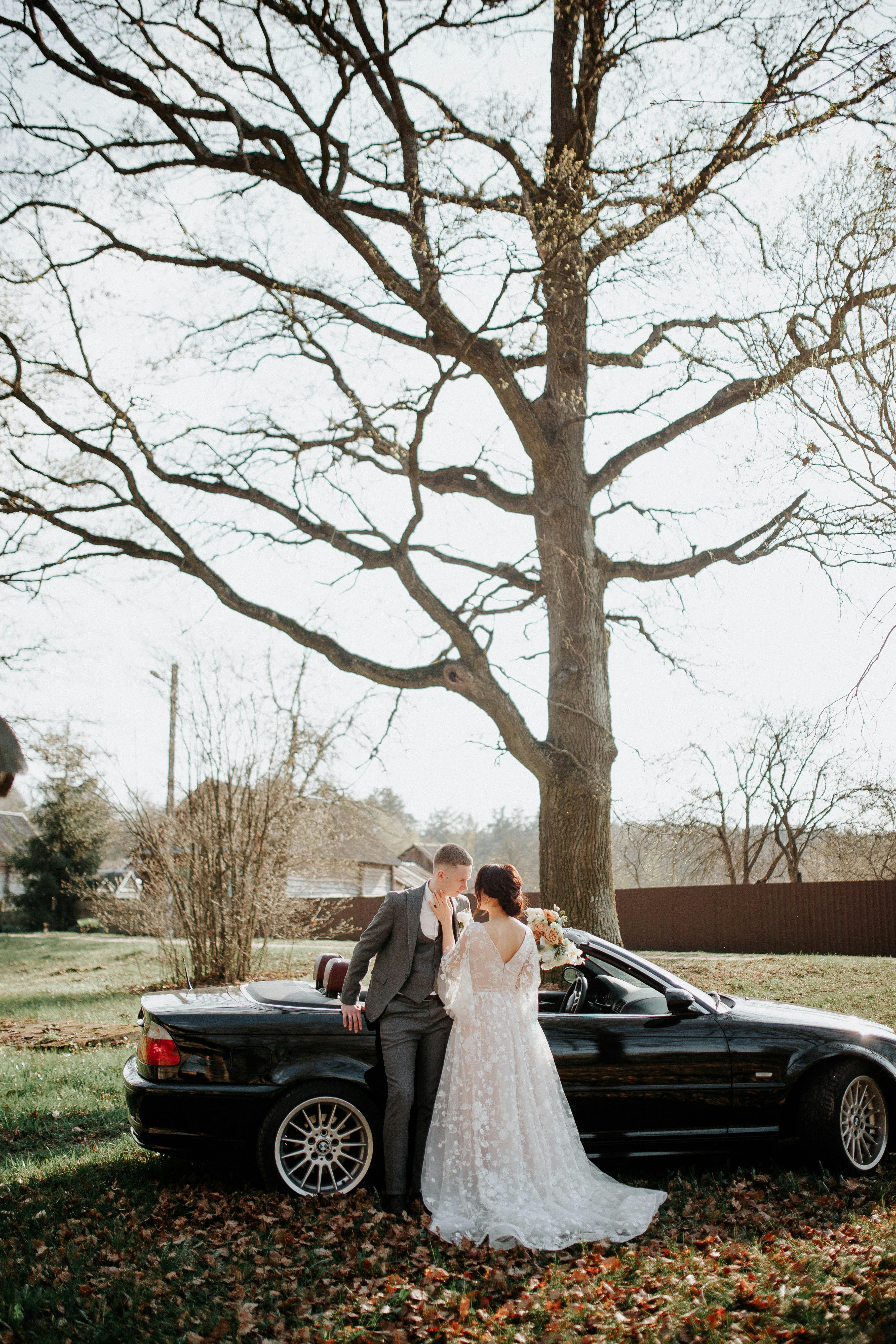 Bride and groom embrace beside a car under a tree on their wedding day.