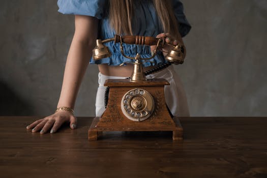 A woman in a blue blouse stands with a vintage wooden rotary phone on a wooden table.