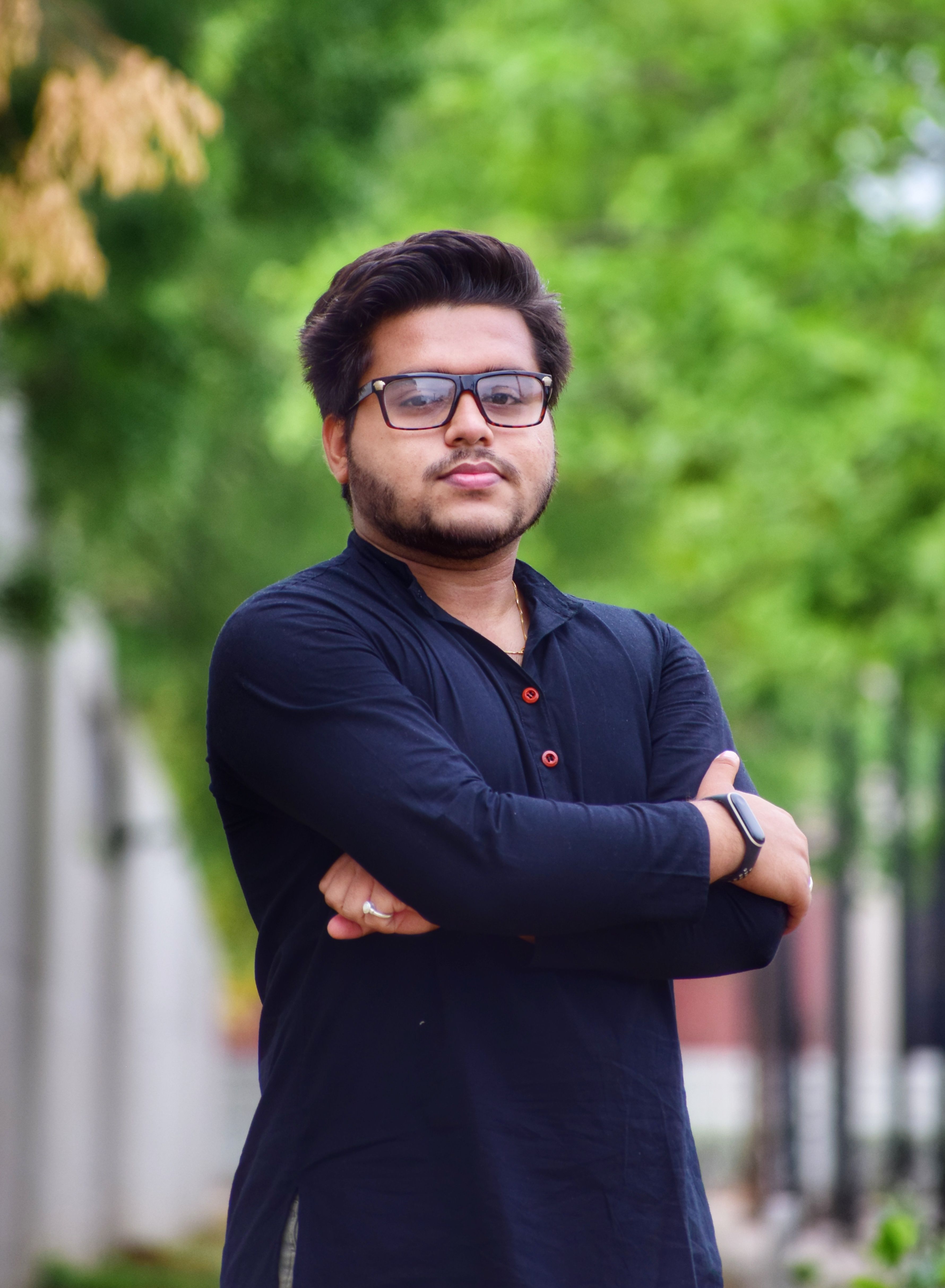 Portrait of a confident young man with eyeglasses posing outdoors in a lush green setting.