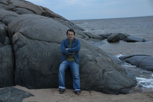 Man leans against rocky coastal shore in denim outfit, with ocean backdrop.