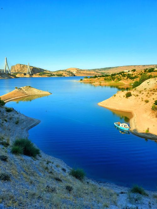 Free Serene blue lake with a small boat and distant bridge, set against rocky hills under a bright sky. Stock Photo
